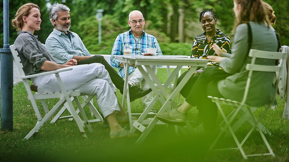 Sechs Personen sitzen um einem Tisch in einem Garten und diskutieren.