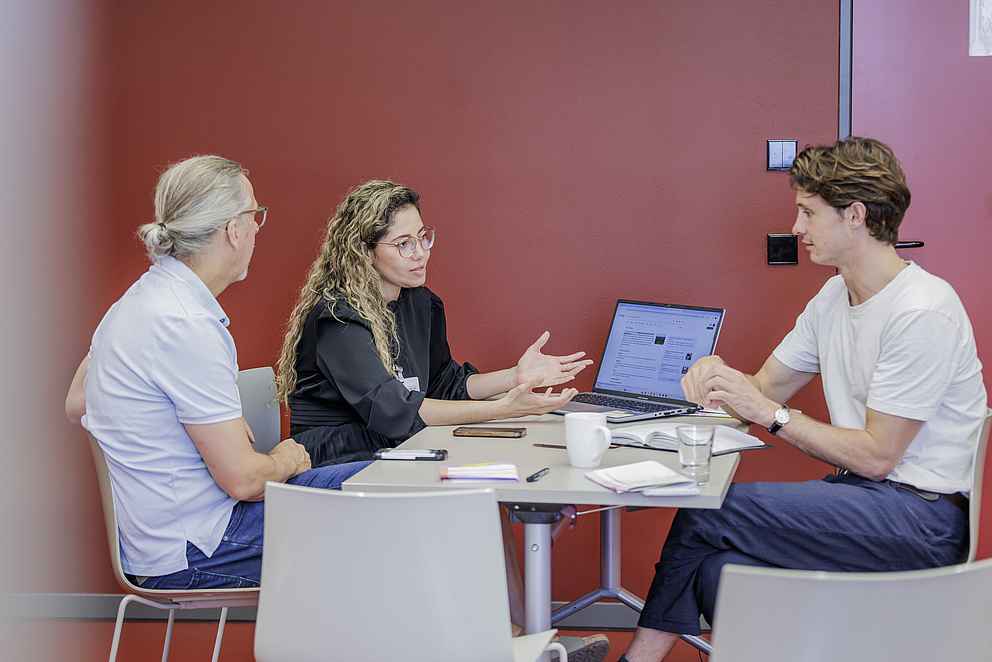 Zwei Männer und eine Frau sitzen um einen Tisch herum. Die Frau erklärt etwas. 