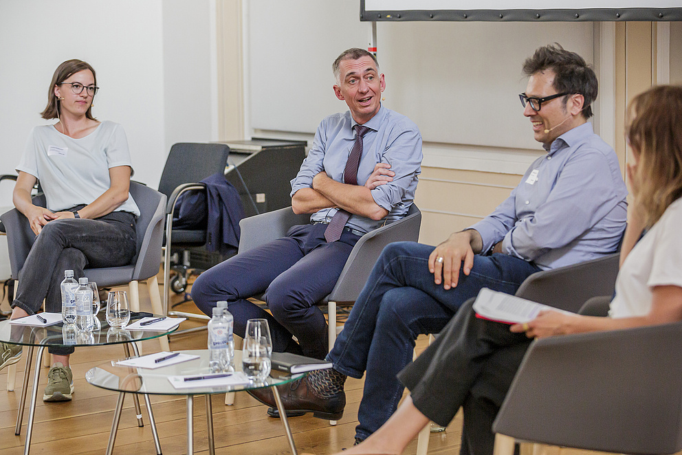 Gezeigt werden vier Personen, die bei einer Paneldiskussion miteinander sprechen.