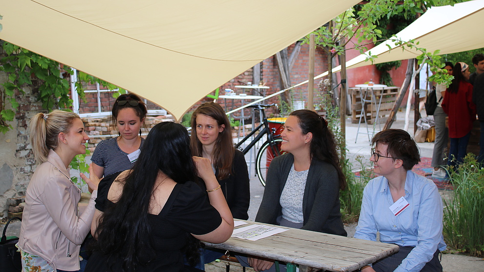 Sechs Personen sitzen an einem Tisch in einem Garten und diskutieren