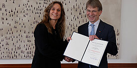 Katja Keul, Staatsministerin im Auswärtigen Amt, steht neben dem Präsidenten der Alexander von Humboldt-Stiftung Hans-Christian Pape. Beide schauen lachend in die Kamera. Sie halten eine Mappe mit einer Urkunde in den Händen.