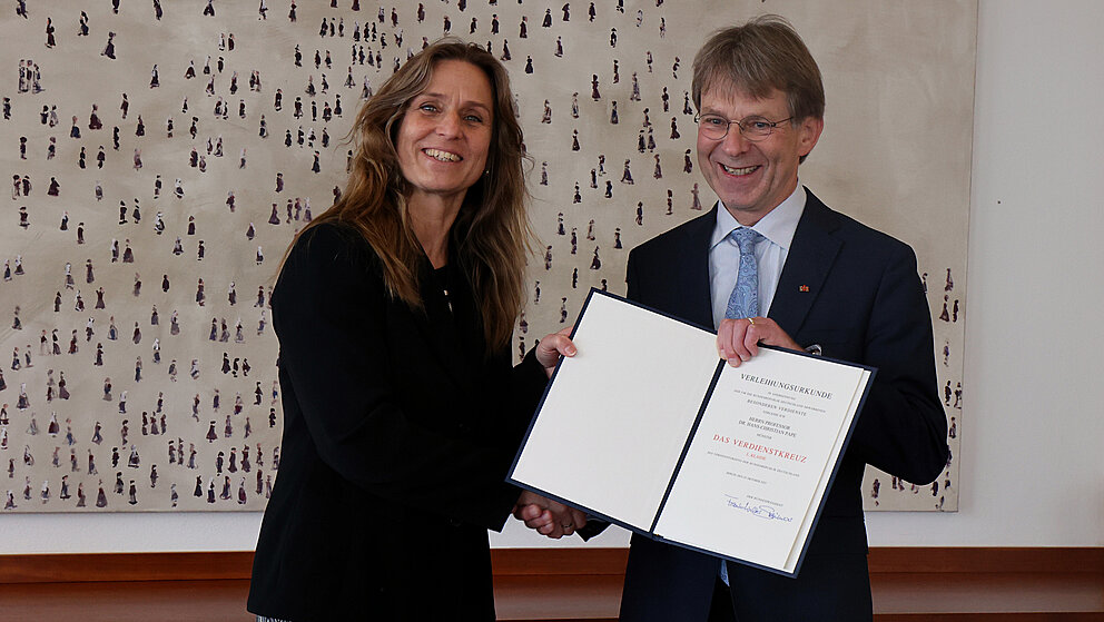 Katja Keul, Staatsministerin im Auswärtigen Amt, steht neben dem Präsidenten der Alexander von Humboldt-Stiftung Hans-Christian Pape. Beide schauen lachend in die Kamera. Sie halten eine Mappe mit einer Urkunde in den Händen.