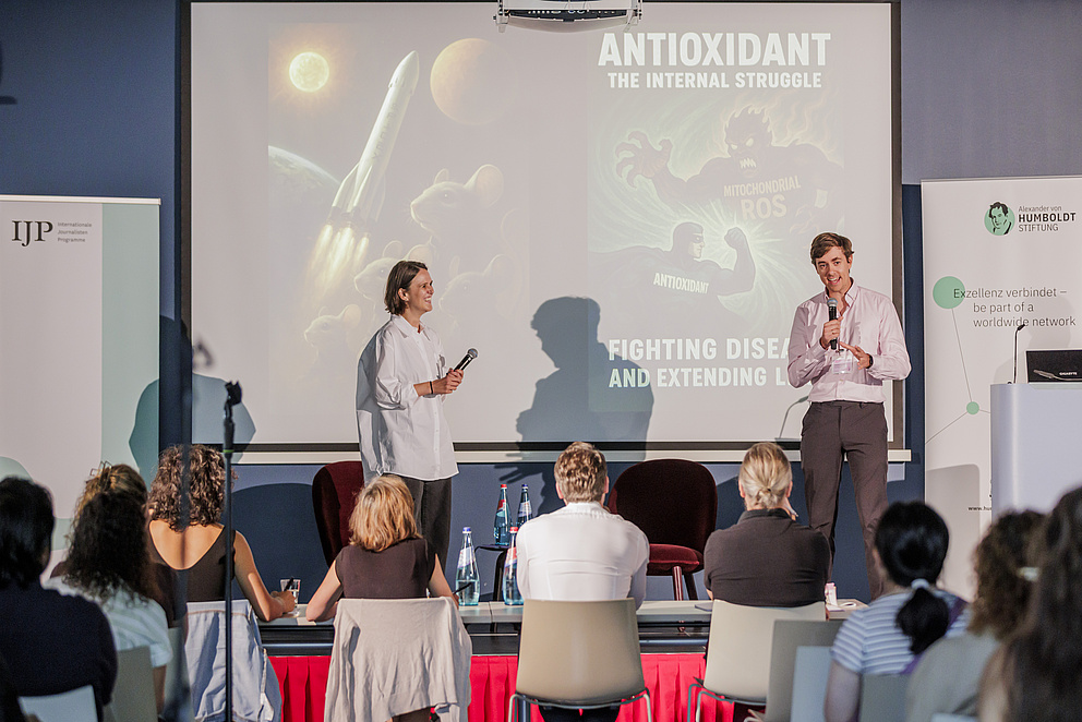 Eine Frau und ein Mann stehen auf einer Bühne und erzählen lachend etwas. Im Hintegrund sieht man eine Präsentation mit dem Text "Antioxidant. The internal struggle."
