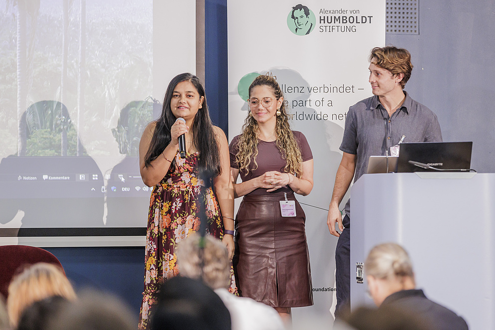 Zwei Frauen und ein Mann stehen auf einer Bühne, vor dem Banner der Humboldt-Stiftung. Die Frau spricht zum Publikum in ein Mikro.