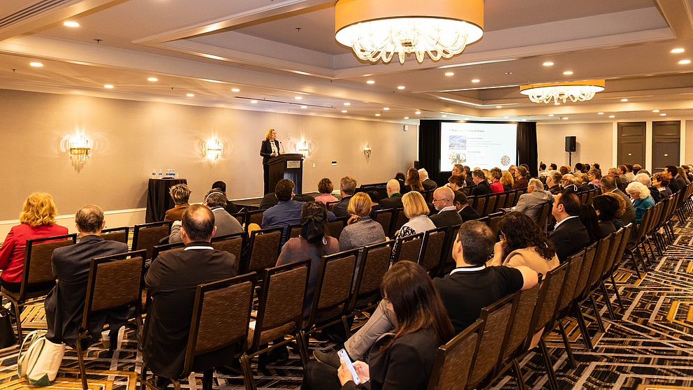 Ein Raum mit einem Rednerpult und ca. 100 Menschen, die auf Stühlen davor sitzen.