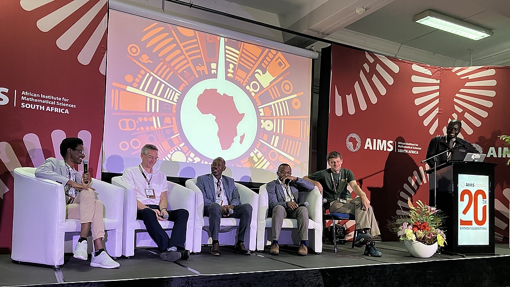 Fünf Männer sitzen auf Sessen auf einer Bühne und unterhalten sich. Einige lachen. Daneben steht ein Mann hinter einem Rednerpult.