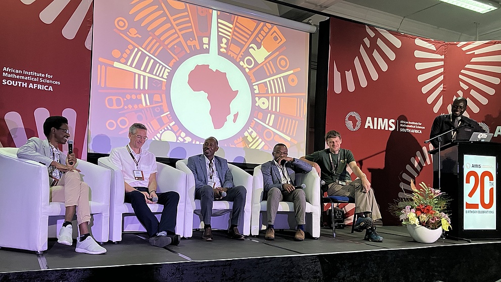 Fünf Männer sitzen auf Sessen auf einer Bühne und unterhalten sich. Einige lachen. Daneben steht ein Mann hinter einem Rednerpult.