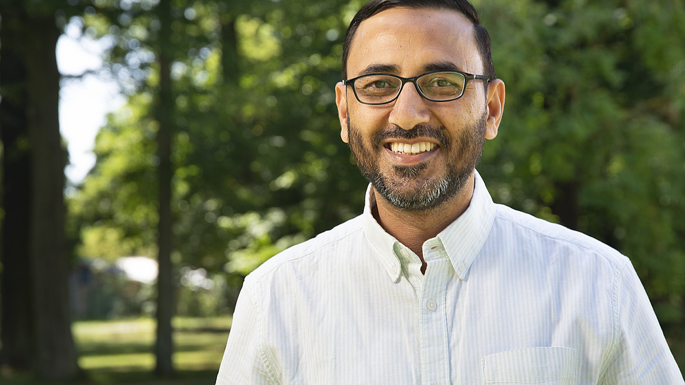 Ein lächelnder Mann mit Brille steht in einem sonnigen Park, umgeben von grünen Bäumen und Rasen.