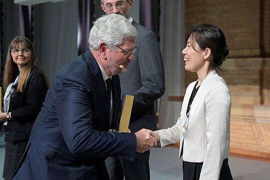 Stiftungspräsident Schlögl schüttelt die Hand von Miki Ebisuya und hält ihre Trophäe