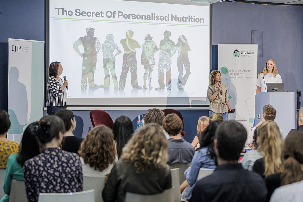 Das Publikum in einem Raum ist von hinten zu sehen. Auf der Bühne stehen drei Frauen, eine von ihnen spricht zum Publikum. Der Text auf der Präsentation im Hintergrund "The secret of personalised nutrition"