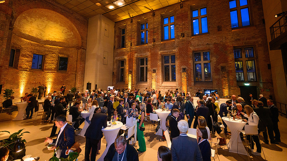Viele Menschen stehen mit Essen und Getränken an Stehtischen in einem hohen, kirchenähnlichen Raum