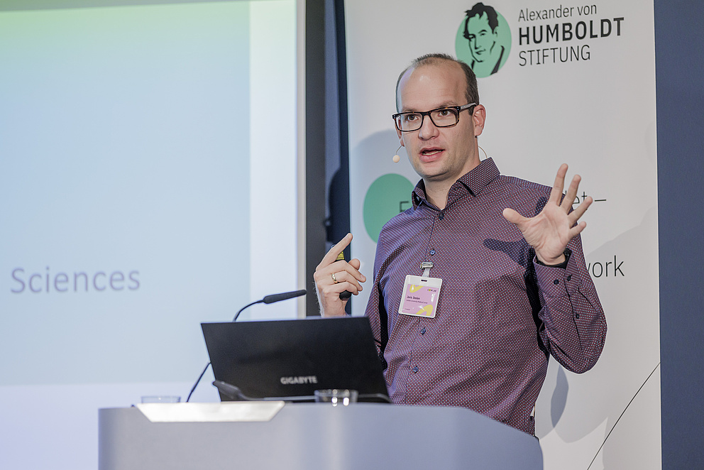 Foto eines Mannes hinter einem Rednerpult, der zum Publikum spricht und gestikuliert.