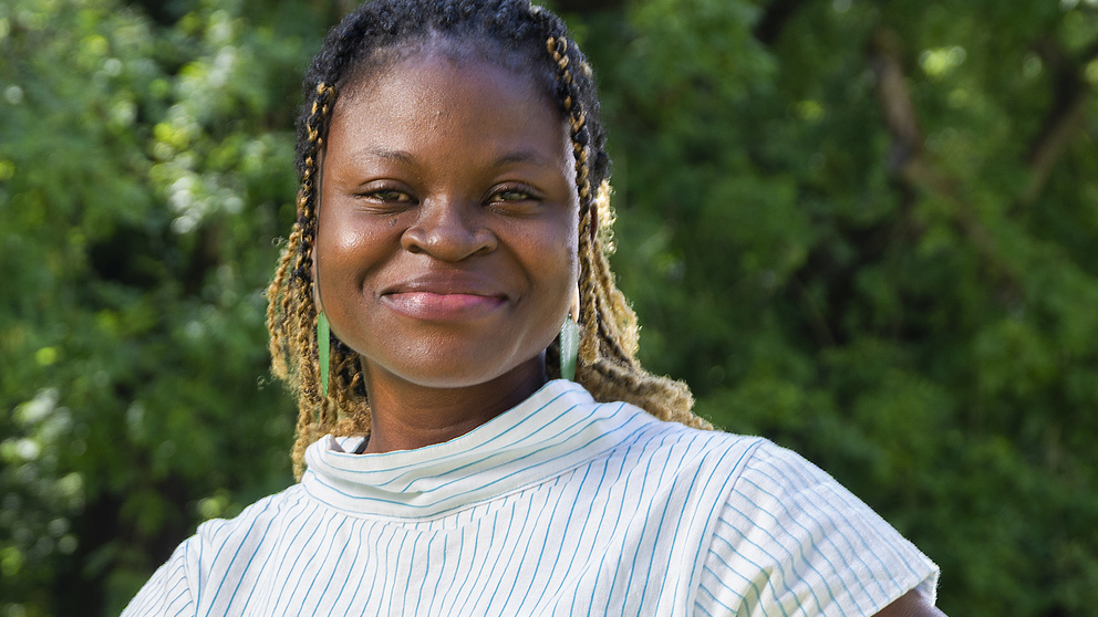 Eine lächelnde Frau mit dunkler Hautfarbe und geflochtenen Haaren steht in einem Park vor grünen Bäumen im Hintergrund.