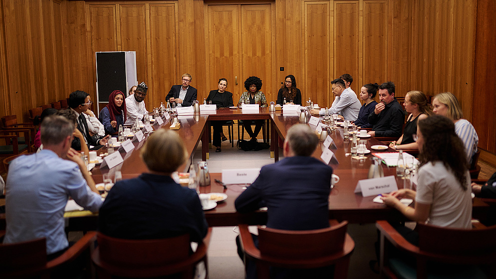 Menschen sitzen an Tischen, die zu einem großen Viereck aufgestellt sind, und diskutieren gemeinsam.