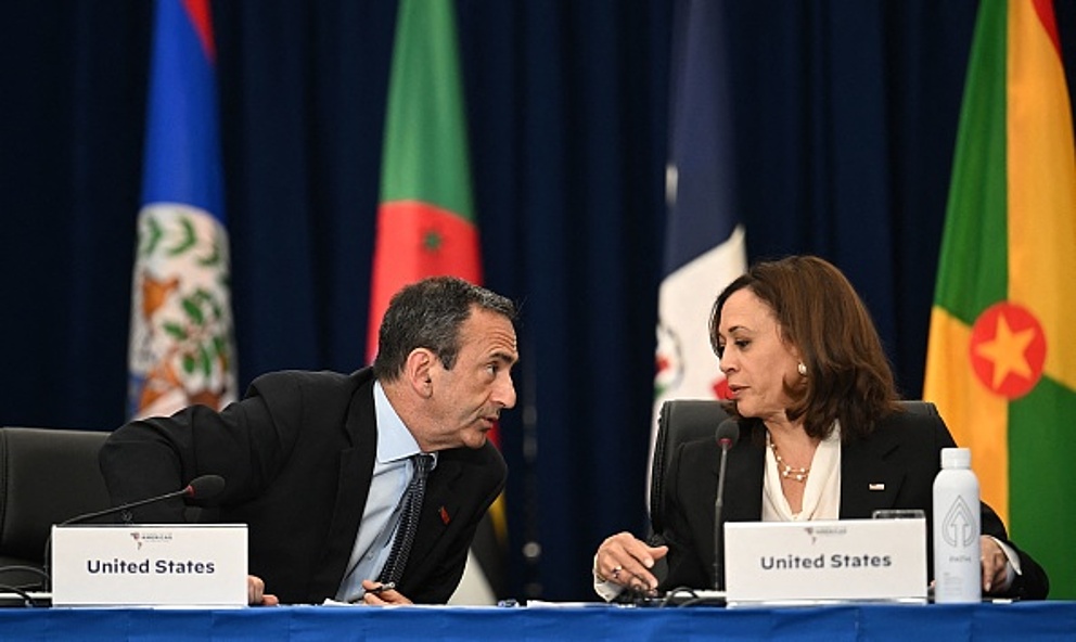 Ein dunkelhaariger Mann und eine dunkelhaarige Frau sitzen nebeneinander hinter einem Tisch. Sie sagen etwas zueinander. Vor ihnen stehen Pappschilder, auf denen jeweils "United States" steht. Es handelt sich um Kamala Harris und Phil Gordon.