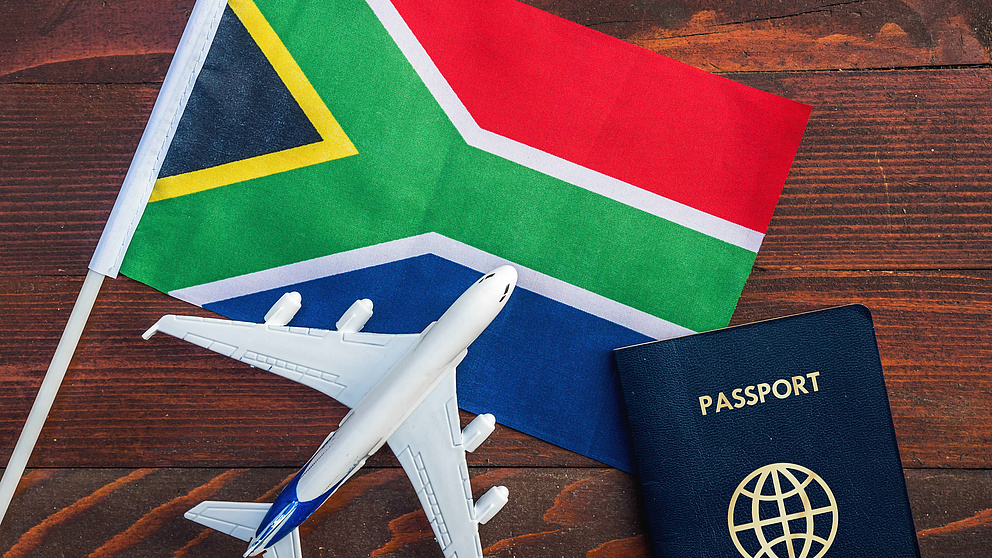 Symbolbild mit Südafrika-Flagge, Modellflugzeug und Reisepass