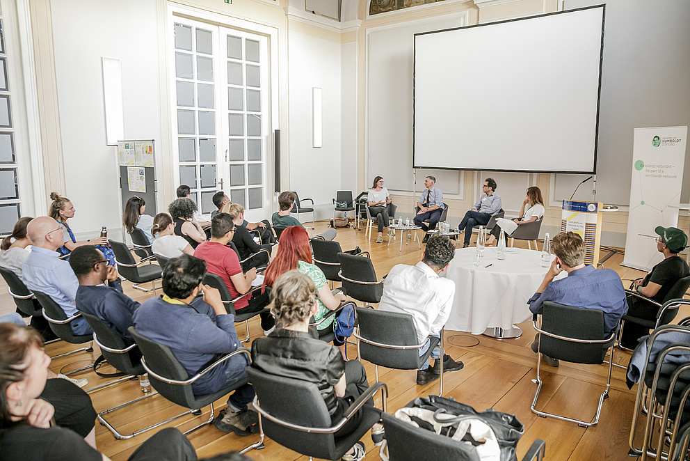 Zu sehen ist eine Menschenmenge auf Stühlen von hinten, die einer Paneldiskussion zuhört.