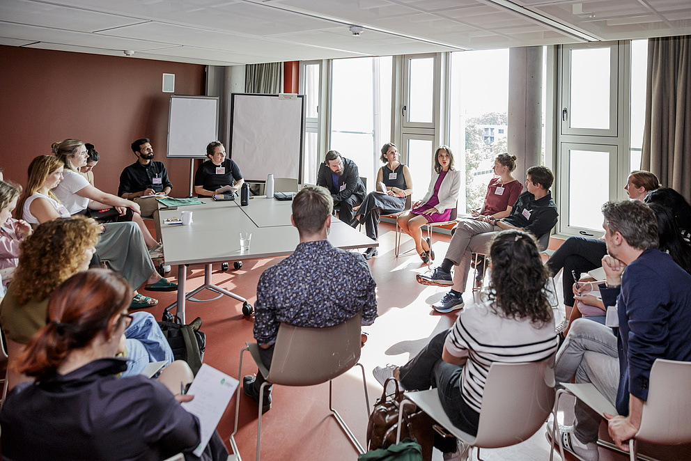 Ein Raum ist zu sehen, in dem Menschen auf Stühlen und Flipcharts zu sehen sind.
