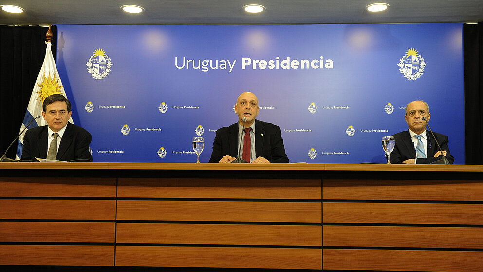 Drei Personen sitzen hinter einem Pult auf einer Pressekonferenz der GACH; in der Mitte: Rafael Radi als Generalkoordinator 