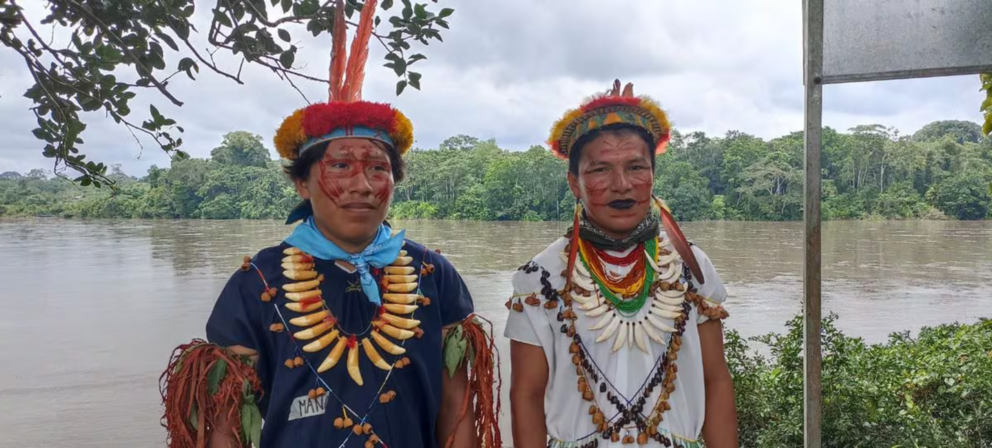 Zwei Mitglieder der Siekopai-Nation in traditioneller Kleidung stehen an einem Fluss mit Regenwald im Hintergrund