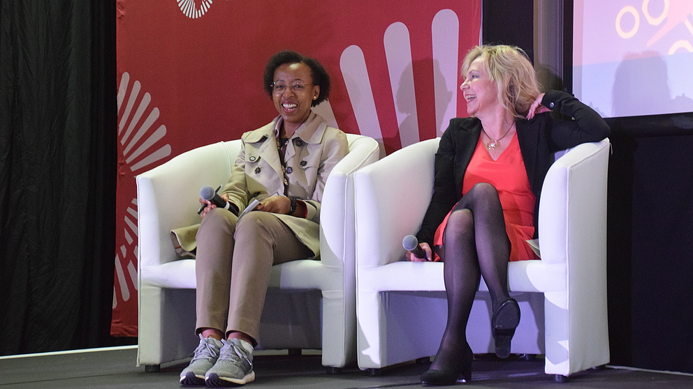 Eine blonde und eine schwarzhaarige Frau sitzen nebeneinander auf einer Bühne auf Sesseln und lachen sich an.