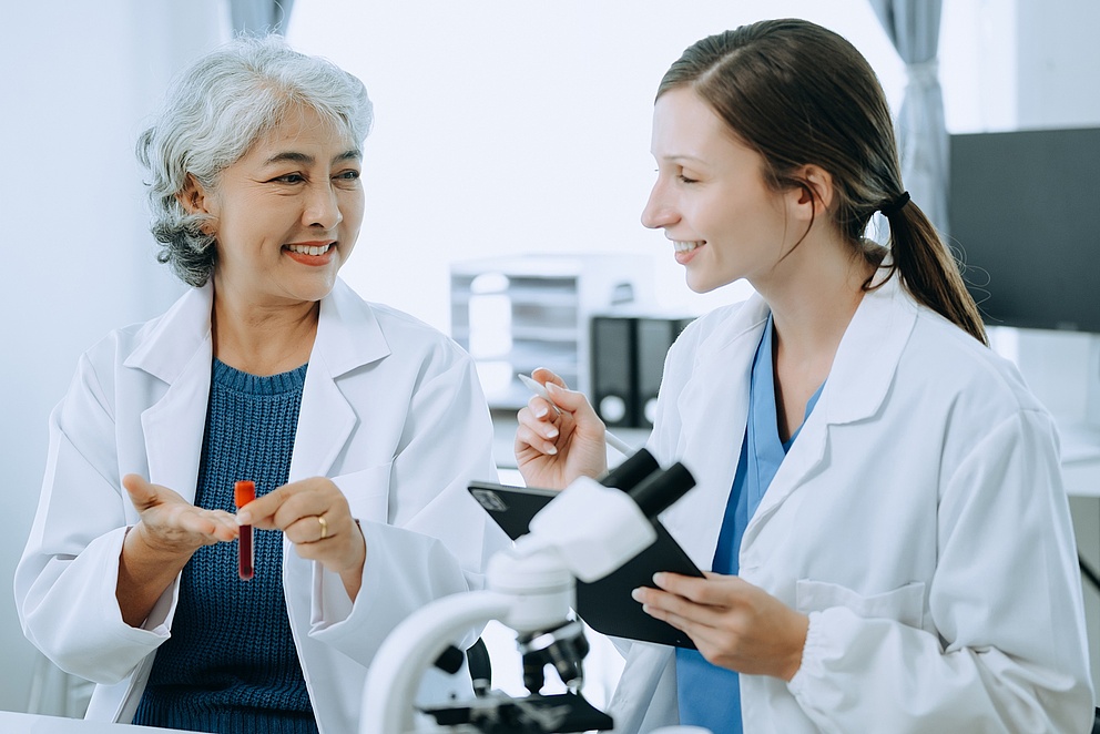 Zwei Wissenschaftlerinnen stehen in einem Labor und lächeln sich an. Beide tragen weiße Laborkittel. Die ältere Wissenschaftlerin hat weiße Haare und hält ein Reagenzglas mit einer Probe in der Hand. Die jüngere hat braune, zusammengebundene Haare und hält ein Tablet sowie einen Stift.