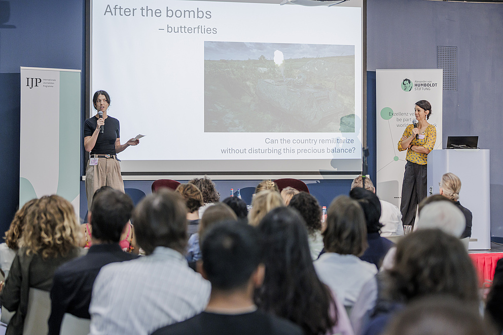 Zwei Frauen stehen auf einer Bühne, die eine spricht zum Publikum. Im Hintergrund ist eine Präsentation zu sehen, mit einem Bild und dem Text "After the bombs - butterflies. Can the country remilitarize witout disturbing this precious balance?"