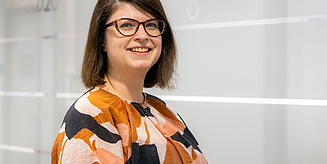 Eine Frau mit Brille, schulterlangen braunen Haaren und einem gemusterten Oberteil lächelt freundlich vor einer modernen, hellen Bürowand.