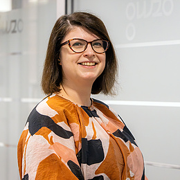 Eine Frau mit Brille, schulterlangen braunen Haaren und einem gemusterten Oberteil lächelt freundlich vor einer modernen, hellen Bürowand.