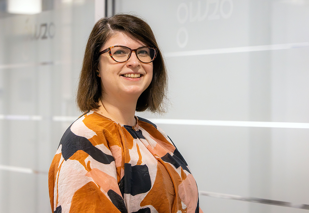 Eine Frau mit Brille, schulterlangen braunen Haaren und einem gemusterten Oberteil lächelt freundlich vor einer modernen, hellen Bürowand.