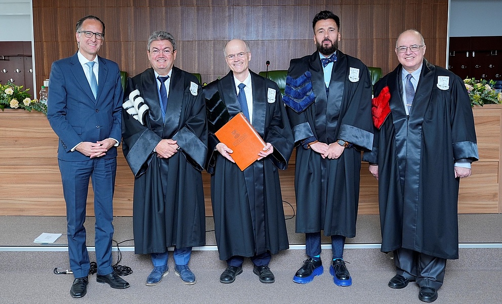 Fünf Männer, vier davon in Roben, stehen nebeneinander und schauen in die Kamera. Der Mann in der Mitte hält eine orangene Mappe in der Hand.