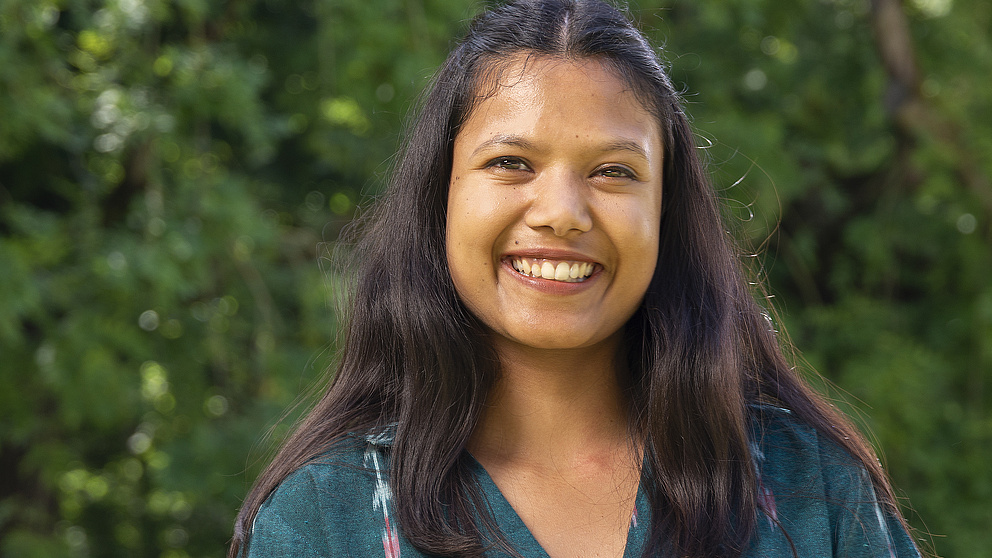 Eine lächelnde junge Frau mit langen, dunklen Haaren steht in einem sonnigen Park vor grünen Bäumen.