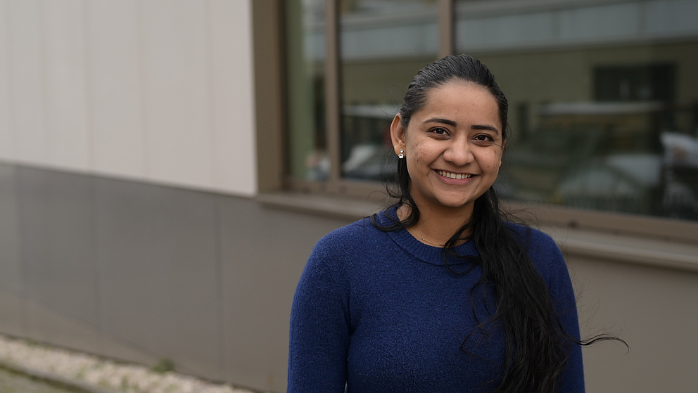 Eine Frau (Sai Kala Kondepudi) mit langen schwarzen Haaren und einem blauen Pullover lächelt in die Kamera. Sie steht vor einem modernen Gebäude mit einer Glasfassade, in der sich ein Teil der Umgebung spiegelt.