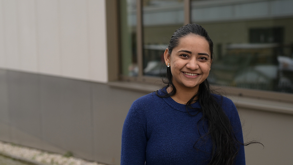Eine Frau (Sai Kala Kondepudi) mit langen schwarzen Haaren und einem blauen Pullover lächelt in die Kamera. Sie steht vor einem modernen Gebäude mit einer Glasfassade, in der sich ein Teil der Umgebung spiegelt.