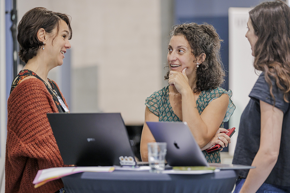 Drei Frauen stehen um einen Tisch herum und sprechen angeregt miteinander.