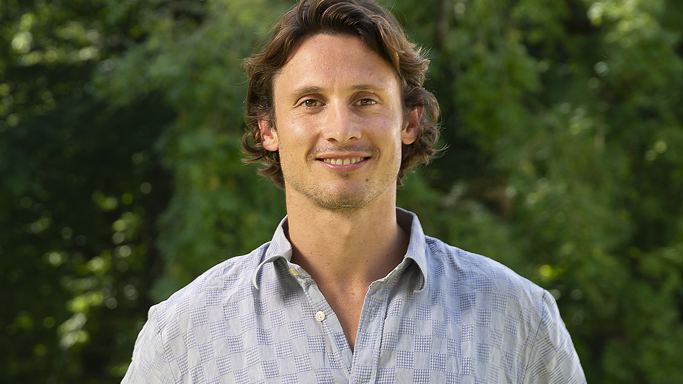 Ein lächelnder junger Mann mit lockigem Haar steht in einem sonnigen Park vor grünen Bäumen.