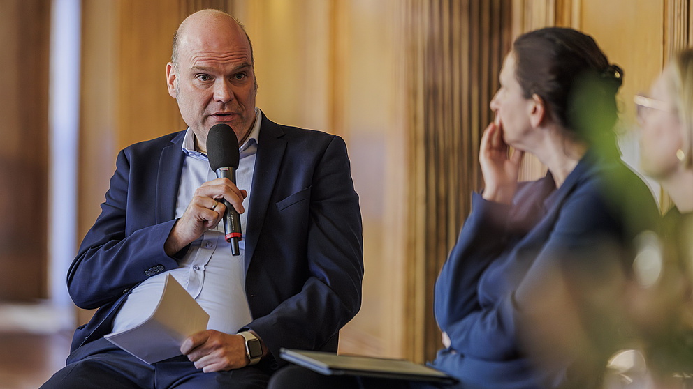 Ein Mann und eine Frau sitzen auf Stühlen auf einer Bühne. Der Mann spricht in ein Mikrofon, die Frau hört zu.