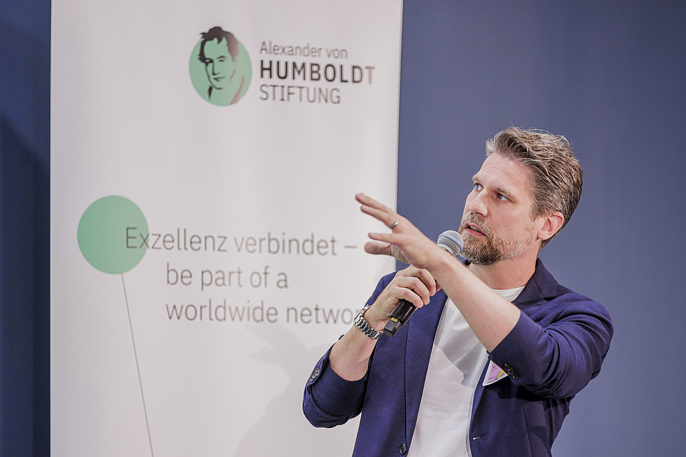 Ein Mann steht auf der Bühne vor dem Banner der Humboldt-Stiftung und zeigt auf etwas. 