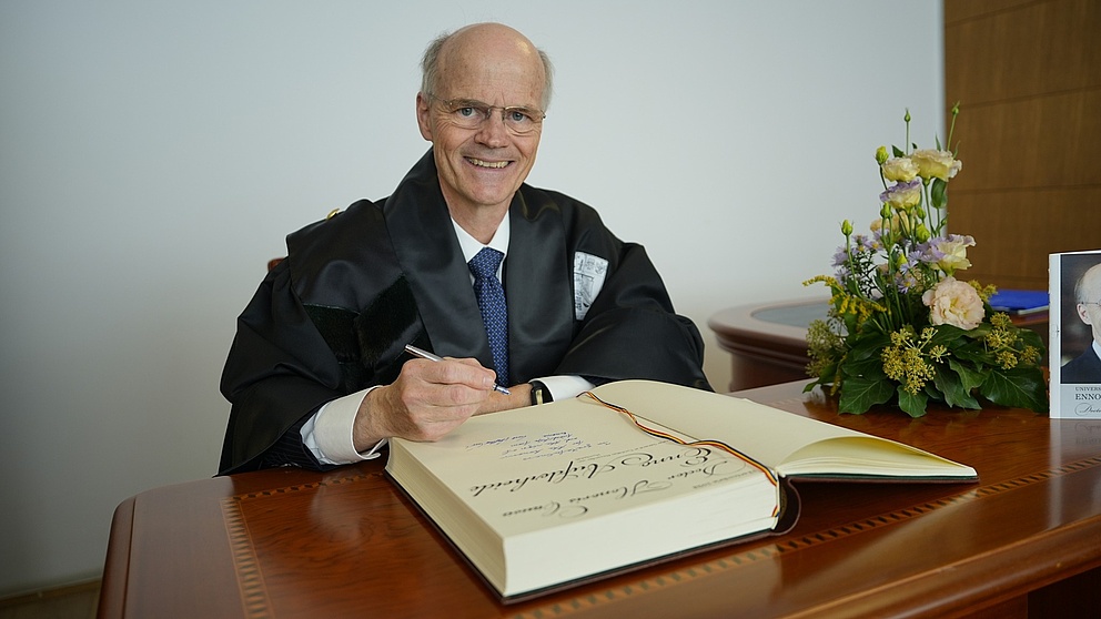 Ein älterer Mann mit Halbglatze sitzt hinter einem Schreibtisch. Er hält einen Stift in der Hand, vor ihm liegt ein großes Buch.