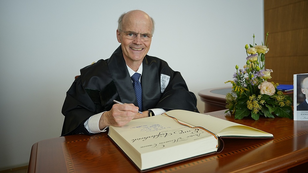 Ein älterer Mann mit Halbglatze sitzt hinter einem Schreibtisch. Er hält einen Stift in der Hand, vor ihm liegt ein großes Buch.