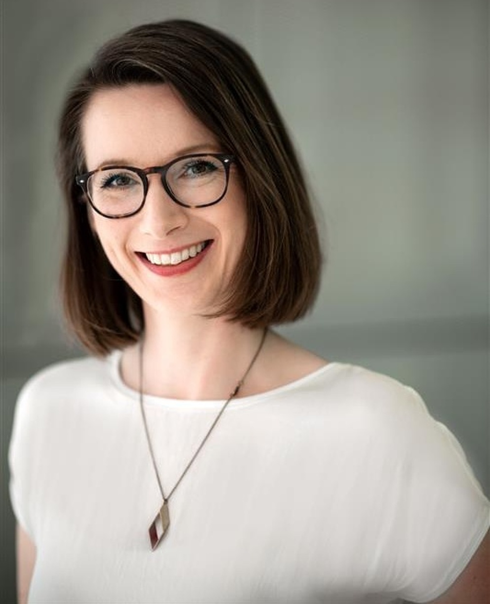 Lächelnde Frau mit boblangem Haar, die eine schwarz-braun gerahmte Brille, eine weiße Bluse und eine einfache Halskette mit Anhänger trägt und direkt in die Kamera schaut.