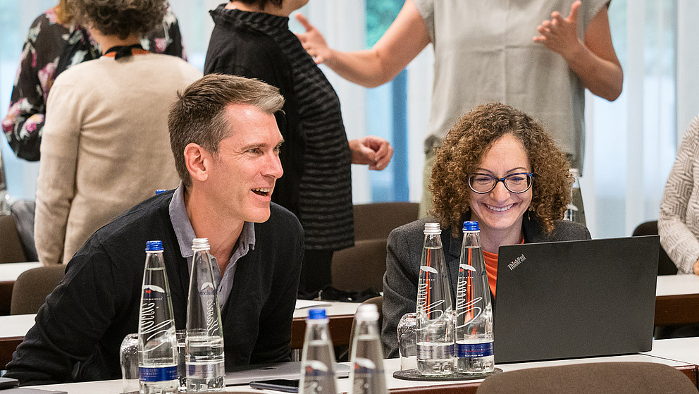 Ein fröhlicher Austausch zwischen einem Mann und einer Frau an einem Tisch, während im Hintergrund andere Teilnehmer aktiv diskutieren.