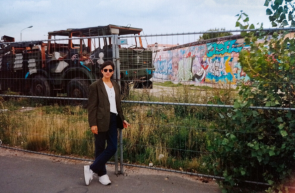 Frau steht vor einem Bauzaun, im Hintergrund die Berliner Mauer, datiert 1994