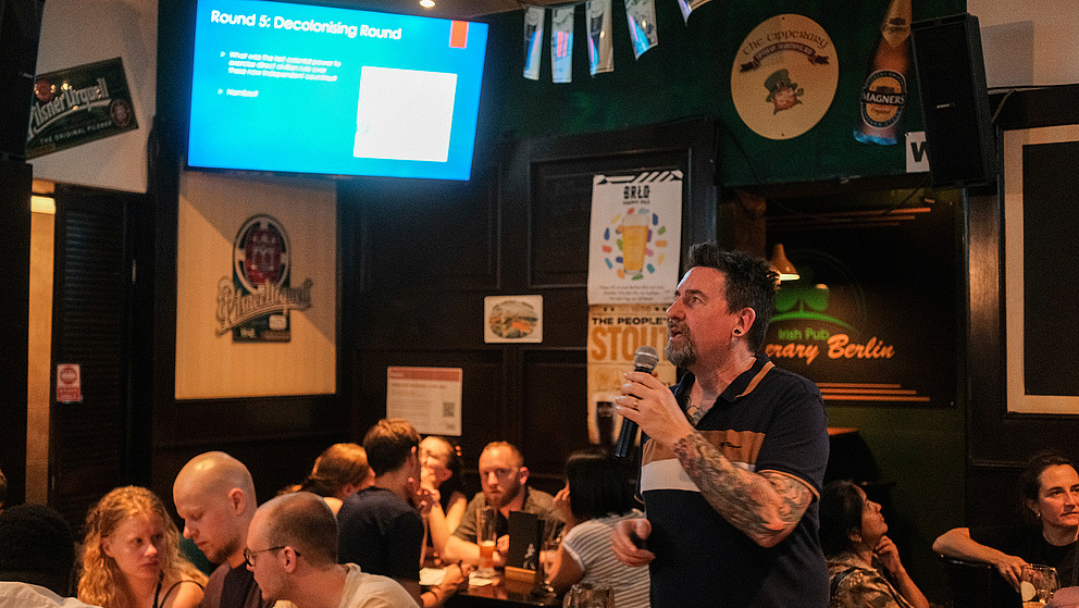 Ein Mann mit einem Mikrofon hin der Hand spricht zu Menschen, die in einem Pub sitzen. Im Hintergrund hängt ein Bildschirm, auf dem ein Text zu sehen ist.