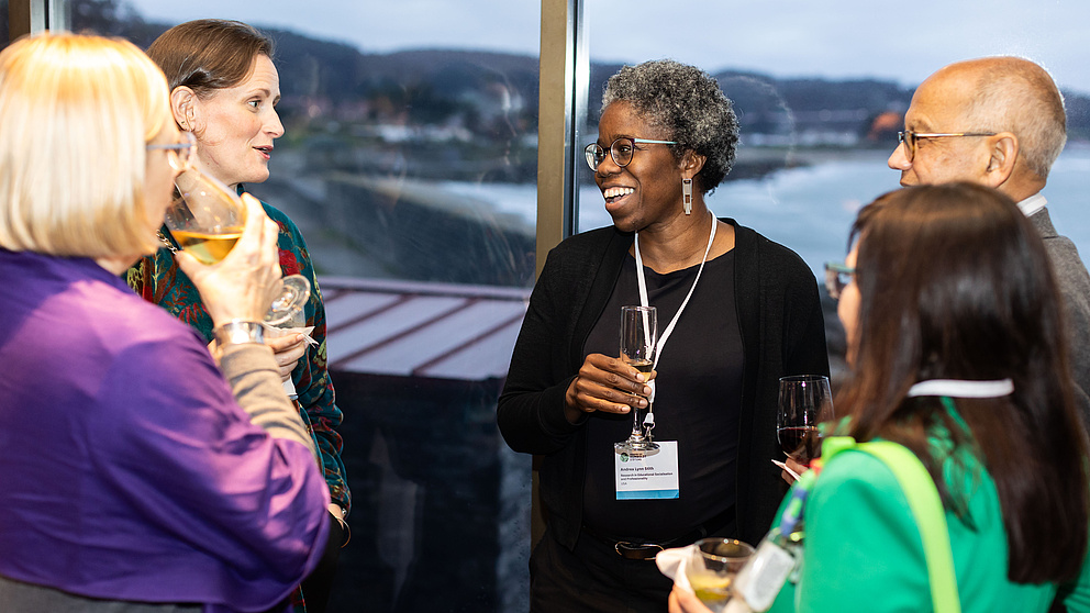 Vier Frauen und ein Mann stehen vor einem Fenster und unterhalten sich lachend. 