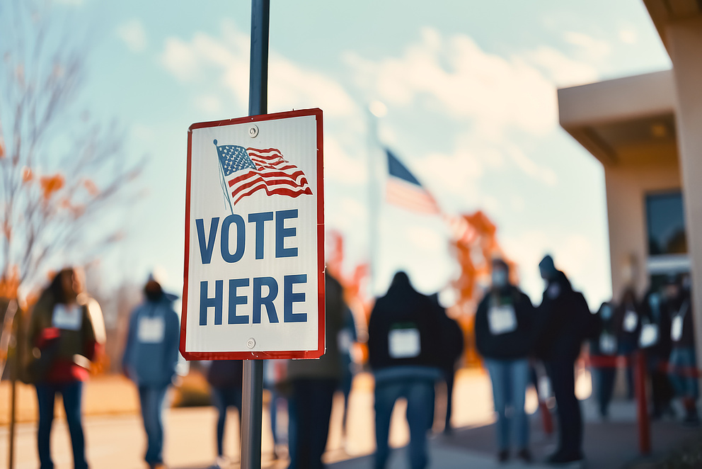 Foto von Wählern, die in einer Schlange vor einem Wahllokal mit dem Schild „Vote Here“ stehen