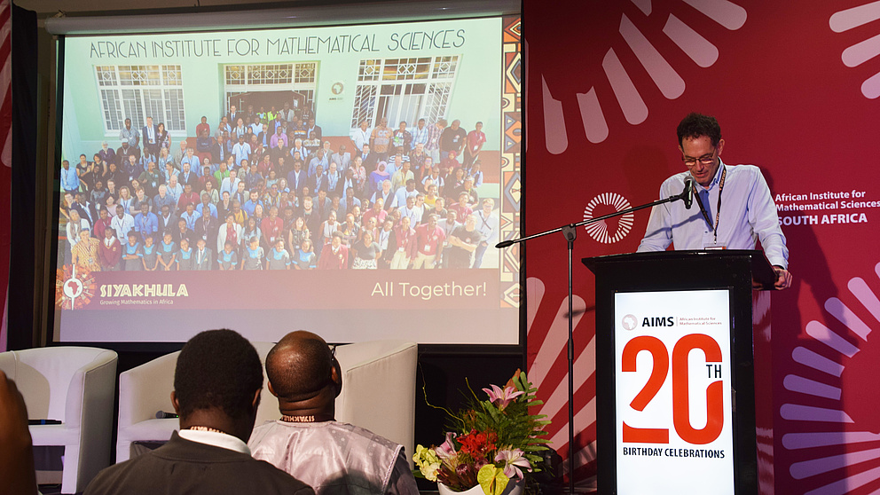 Ein Mann steht hinter einem Rednerpult, neben ihm läuft eine PowerPoint-Präsentation auf einer Leinwand