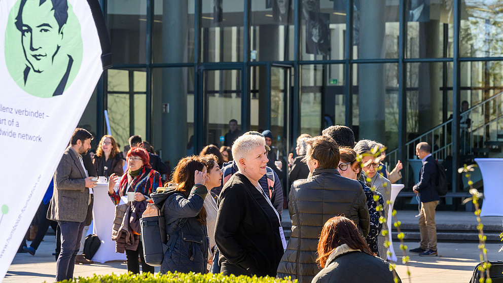 Menschen stehen vor der FU Berlin und unterhalten sich. Eine Fahne mit dem Humboldt-Logo ist ausserdem zu sehen.