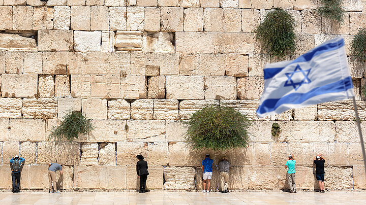 Die Klagemauer in Jerusalem mit betenden Menschen. Rechts im Vordergrund unscharf eine Israel-Flagge
