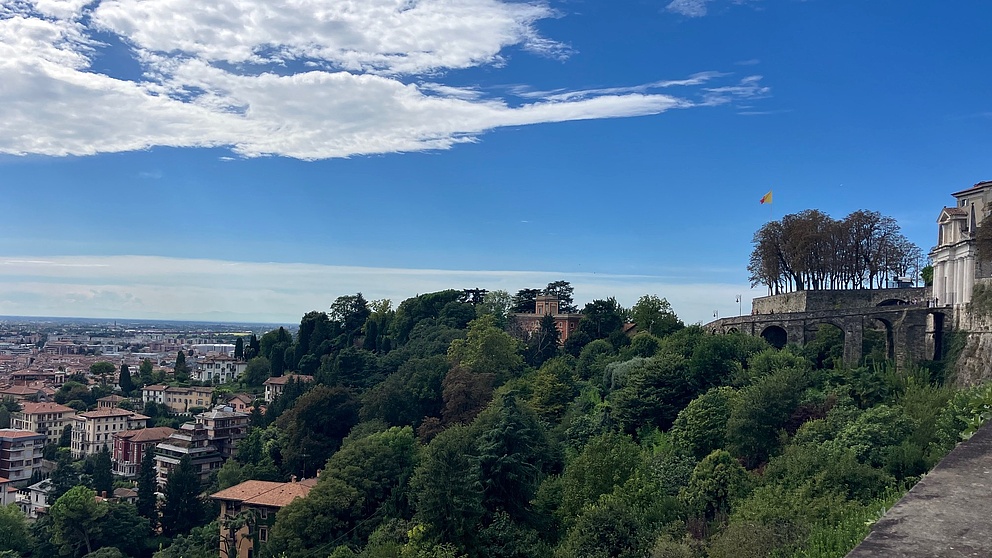 The photo shows a panoramic view of the city of Bergamo under a bright blue sky with a few clouds. Numerous houses and green trees characterize the cityscape, on the right you can see a historic building.